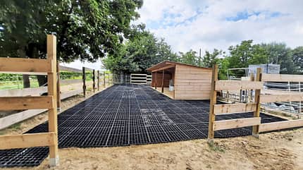 ridcon, Paddockbau, Paddockboden, Kunststoffgitterplatte, Bodenbefestigung, Weideunterstand, Pferdehaltung, Reitanlage, Holzzaun, Auslauf Das Foto zeigt einen Paddockbau mit ProGrid-Elementen von ridcon. Die verlegten schwarzen Kunststoff‑Gitterplatten dienen als Bodenbefestigung auf einem Sandpaddock. Zwischen den Holzzäunen führt die befestigte Auslauffläche zu einem hölzernen Weideunterstand; rechts sind zusätzliche Paddock-Elemente und Panels zu sehen.