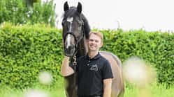 Raphael Netz, Pferdeporträt, Reiterporträt, Mensch-und-Tier, Halfter, Reitsport, Tierfotografie, Outdoor-Porträt, Grünanlage, Sommer, 2026 Raphael Netz neben einem dunkelbraunen Pferd mit schmaler Stirnblässe; er hält das Tier am Halfter vor einer dichten grünen Hecke. Es handelt sich um ein natürlich beleuchtetes Outdoor‑Porträt mit geringer Schärfentiefe, unscharfen Vordergrundelementen und klar erkennbarem Halfter.