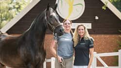 Vicky Telgen, Ariane Telgen, Hof Eicheneck, Pferdeporträt, Reitsport, Pferd-Mensch-Porträt, Stallhof, Halfter, Warmblut, Tierfotografie, Cavallo Das Foto Vicky Telgen und ihre Mutter Ariane Telgen mit Pferd auf Hof Eicheneck vor einem Stallgebäude mit Logo. Das dunkelbraune Warmblut steht im Halfter, eine Führleine ist sichtbar; weiße Zaunelemente und die Holzfassade bilden den Hintergrund.