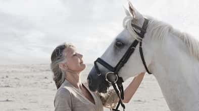 Eine grauhaarige Frau mit einem Schimmel am Strand.