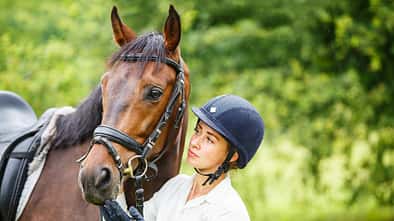 Eine junge Frau mit Reithelm hält ihr aufgetrenstes Pferd im Grünen.
