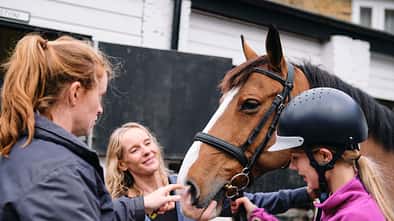 Mother and instructor preparing young girl for horseback riding lesson