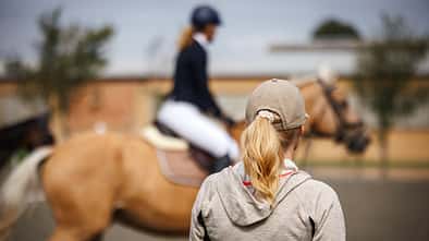Eine Frau von hinten mit Blick in Richtung einer Turnierreiterin auf dem Abreiteplatz