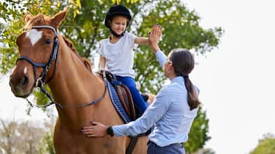 Ein junges Mädchen auf dem Pferd klatscht sich mit der Trainerin am Boden ab