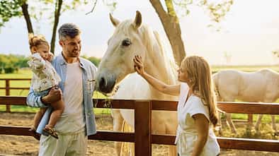 Eine Familie mit einem kleinen Mädchen interagiert mit einem weißen Pferd auf einem Bauernhof bei Sonnenuntergang. Die Szene zeigt eine emotionale Bindung zwischen den Familienmitgliedern und dem Tier in ländlicher Umgebung.