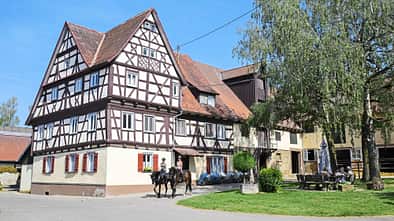 Das Foto zeigt den Birkhof mit einem großen historischen Fachwerkhaus und angrenzenden Stallgebäuden. Im Hof reiten zwei Personen auf Pferden vorbei, rechts sitzen Menschen an einem Holztisch unter Bäumen; geöffnete Stall- und Scheunentore sind sichtbar.