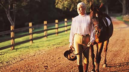 Ältere attraktive Frau mit Helm in der Hand führt ihr Pferd vorbei an den Weiden zur Reitbahn.