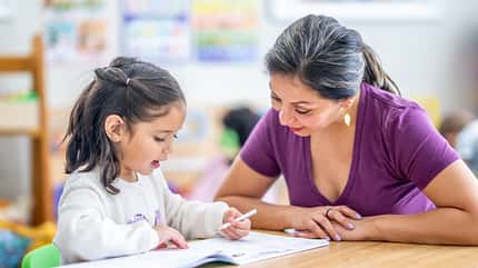 Ein Mädchen sitzt mit ihrer Lehrerin am Tisch und lernt schreiben
