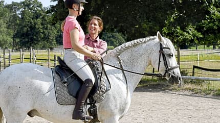 Reitunterricht, Pferd, Reiterin, Trainerin, Dressur, Reitplatz, Sitzschulung, Pferdesport, Helm, weißer Schimmel Eine Reiterin sitzt auf einem weißen Pferd, während eine Trainerin neben ihr steht und Anweisungen gibt. Die Szene spielt auf einem Reitplatz im Freien. Das Pferd ist gesattelt und trägt eine Trense, die Reiterin trägt Reitbekleidung und einen Helm.