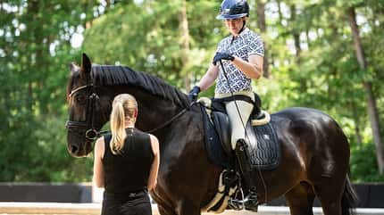 Reitsport, Dressur, Pferd, Reiterin, Trainerin, Helm, Warendorf, Trainertag, Cavallo, Ridingtogether, Katharina Hemmer, Gianna Regenbrecht Para-Reiterin Gianna Regenbrecht im Dressursattel, Katharina Hemmer steht daneben auf dem Reitplatz, Wald im Hintergrund.
