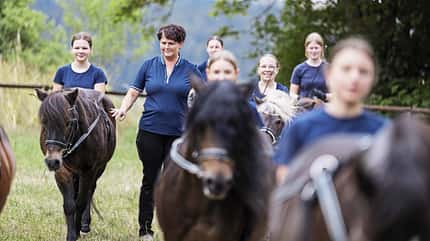 Ponyreiten, Langzügel, Bodenarbeit, Reitunterricht, Reitschule, Pferdesport, Pony, Gruppe, Weide, Freizeitaktivität, Reitlehrerin Das Foto zeigt eine Gruppe von Jugendlichen mit Ponys am Langzügel, angeleitet von einer Reitlehrerin, die in der Mitte der Wiese läuft. Im Hintergrund sind Bäume und eine hügelige Landschaft erkennbar.
