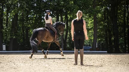 Eine Trainingseinheit im Dressurviereck: Anna Den reitet ihr dunkelbraunes Pferd, während Katharina Hemmer vom Boden aus beobachtet und anleitet.