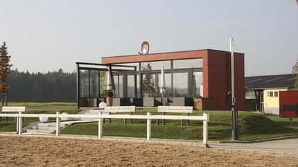 Hippocase, Reiterstübchen, Richterhäuschen, Anbau, Schlosser Stallbau HippoCASE-Bau von Schlosser mit roten Außenwänden und Glasfassade vorne am Rande eines Dressurvierecks.
