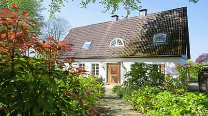 Wohnhaus, Einfamilienhaus, Satteldach, Dachziegel, Dachfenster, Eingangstür, Vorgarten, Gartenweg, Architektur, Immobilie, Gut Scheidt, Angemeer Das Bild zeigt ein Wohnhaus mit Satteldach und weißer Fassade. Sichtbar sind ein Ziegeldach mit Moosbewuchs, zwei Dachfenster, ein halbkreisförmiges Giebelfenster, zwei Schornsteine sowie eine hölzerne Eingangstür; der Zugang führt über einen gepflasterten Gartenweg.