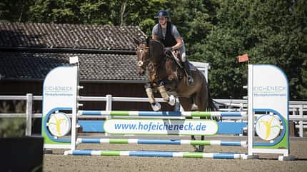 Vicky Telgen, Ariane Telgen, Hof Eicheneck, Springreiten, Hindernisparcours, Reitsport, Springpferd, Reithelm, Schutzweste, Gamaschen, Außenreitplatz, Stangenhindernis, Pferd Das Foto zeigt Vicky auf einem Springpferd beim Überwinden eines farbigen Stangenhindernisses in einem Außenreitplatz. Zu sehen sind Reithelm und Schutzweste, das Pferd trägt Gamaschen; im Vordergrund steht ein mit Werbung versehenes Hindernis („hofeicheneck.de“). Im Hintergrund sind Reitplatzumzäunung und Bäume erkennbar.