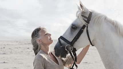 Eine grauhaarige Frau mit einem Schimmel am Strand.