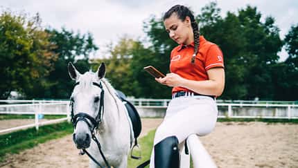 Ein Mädchen sitzt auf der Reitplatz-Umrandung und hält ein Smartphone in der Hand. Ihr Pferd steht daneben.