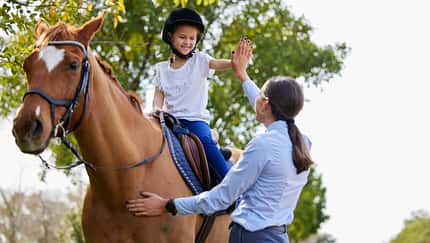 Ein junges Mädchen auf dem Pferd klatscht sich mit der Trainerin am Boden ab