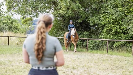 Reitunterricht, Reitstunde, Reiterin, Pferd, Reitplatz, Reithelm, Sattel, Pferdesport, Naturkulisse Das Foto zeigt eine Reiterin mit Reithelm auf einem Haflinger auf einem umzäunten Reitplatz. Im unscharfen Vordergrund steht eine Reitlehrerin, während Bäume und Wiese den natürlichen Hintergrund bilden.