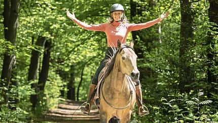 Featherlight Horsemanship, Pferdetraining, Geländereiten, Reithelm, Waldweg, Niederlande, Horsemanship, Freizeitreiten, Reiterin, Pferd, Yvette Blokesch Das Foto zeigt eine Reiterin mit Reithelm auf einem Pferd beim Ausritt auf einem schattigen Waldweg, die Arme seitlich ausgestreckt.