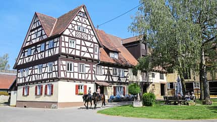 Birkhof, Reiterhof, Fachwerkhaus, Bauernhof, Pferdesport, Pferd, Hofanlage, Stallgebäude, Ländliche Architektur, Ausritt Das Foto zeigt den Birkhof mit einem großen historischen Fachwerkhaus und angrenzenden Stallgebäuden. Im Hof reiten zwei Personen auf Pferden vorbei, rechts sitzen Menschen an einem Holztisch unter Bäumen; geöffnete Stall- und Scheunentore sind sichtbar.