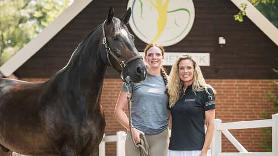 Das Foto Vicky Telgen und ihre Mutter Ariane Telgen mit Pferd auf Hof Eicheneck vor einem Stallgebäude mit Logo. Das dunkelbraune Warmblut steht im Halfter, eine Führleine ist sichtbar; weiße Zaunelemente und die Holzfassade bilden den Hintergrund.