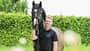 Raphael Netz, Pferdeporträt, Reiterporträt, Mensch-und-Tier, Halfter, Reitsport, Tierfotografie, Outdoor-Porträt, Grünanlage, Sommer, 2026 Raphael Netz neben einem dunkelbraunen Pferd mit schmaler Stirnblässe; er hält das Tier am Halfter vor einer dichten grünen Hecke. Es handelt sich um ein natürlich beleuchtetes Outdoor‑Porträt mit geringer Schärfentiefe, unscharfen Vordergrundelementen und klar erkennbarem Halfter.