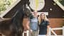 Vicky Telgen, Ariane Telgen, Hof Eicheneck, Pferdeporträt, Reitsport, Pferd-Mensch-Porträt, Stallhof, Halfter, Warmblut, Tierfotografie, Cavallo Das Foto Vicky Telgen und ihre Mutter Ariane Telgen mit Pferd auf Hof Eicheneck vor einem Stallgebäude mit Logo. Das dunkelbraune Warmblut steht im Halfter, eine Führleine ist sichtbar; weiße Zaunelemente und die Holzfassade bilden den Hintergrund.