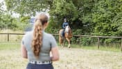 Das Foto zeigt eine Reiterin mit Reithelm auf einem Haflinger auf einem umzäunten Reitplatz. Im unscharfen Vordergrund steht eine Reitlehrerin, während Bäume und Wiese den natürlichen Hintergrund bilden.