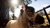 Im Vordergrund ist ein Pony zu sehen, dass von einer lachenden Frau im Hintergrund gerade am Rücken gestreichelt wird. 
