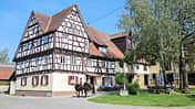 Das Foto zeigt den Birkhof mit einem großen historischen Fachwerkhaus und angrenzenden Stallgebäuden. Im Hof reiten zwei Personen auf Pferden vorbei, rechts sitzen Menschen an einem Holztisch unter Bäumen; geöffnete Stall- und Scheunentore sind sichtbar.